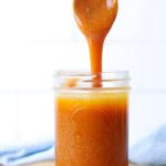 A jar of caramel sauce sitting on a piece of parchment paper. There is a spoon drizzling the caramel into the mason jar.
