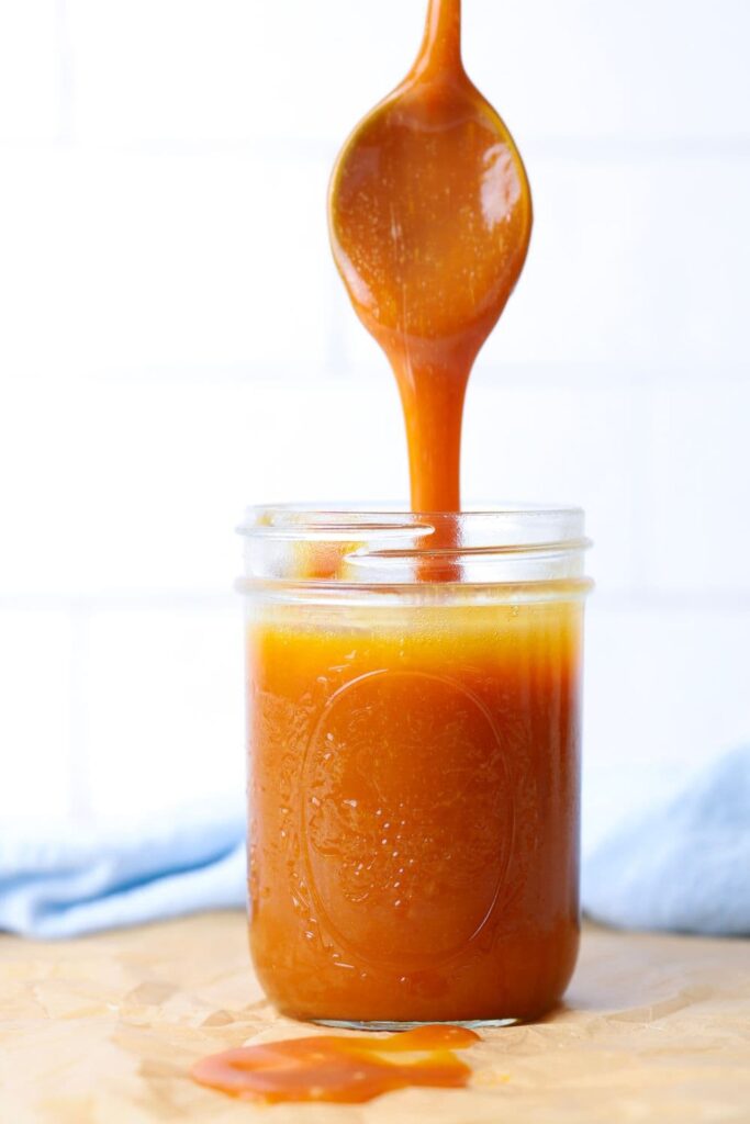 A jar of caramel sauce sitting on a piece of parchment paper. There is a spoon drizzling the caramel into the mason jar.