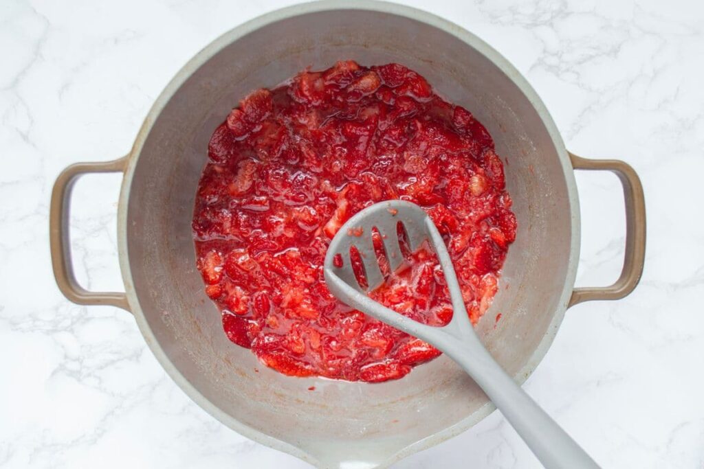 A large handled pot containing mashed red strawberries. There is a potato masher in the pot.