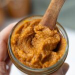 A jar of pumpkin butter being held by a hand. There is a wooden spoon in the top of the jar.