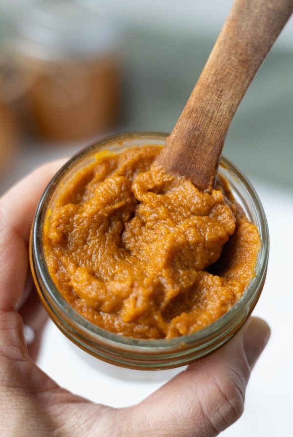 A jar of pumpkin butter being held by a hand. There is a wooden spoon in the top of the jar.