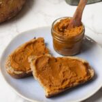 2 slices of sourdough bread spread with pumpkin butter. The jar of pumpkin butter is sitting on the plate as well, with the wooden spoon in the jar.