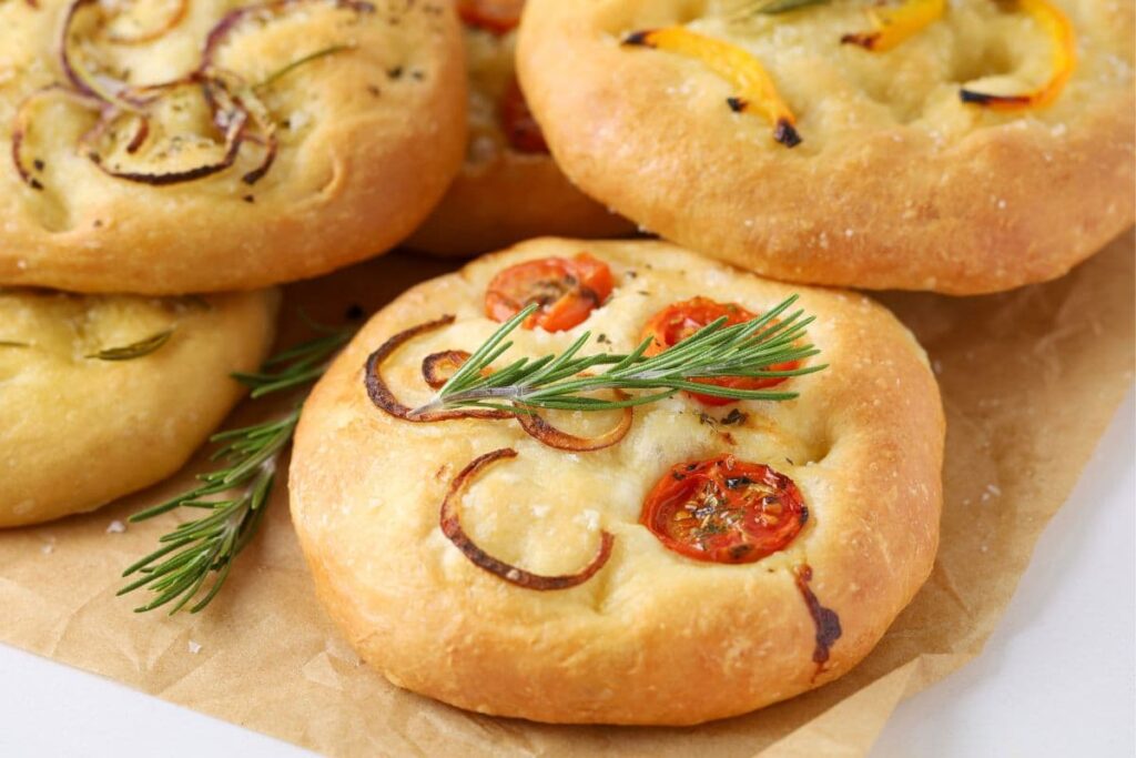 Close up of a sourdough focaccia roll with cherry tomatoes, onions and a sprig of fresh rosemary on top.