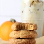 Four sourdough pumpkin snickerdoodle cookies stacked on parchment paper with a coffee drink in background.