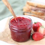 A small jar of strawberry jam sitting on a white plate alongside some fresh strawberries. There is a wooden spoon in the jar and some sourdough in the background.