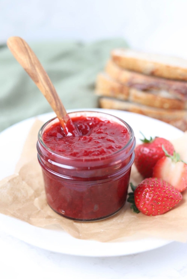 A small jar of strawberry jam sitting on a white plate alongside some fresh strawberries. There is a wooden spoon in the jar and some sourdough in the background.