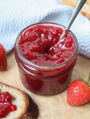 A small jar of strawberry jam with a spoon in it. There are fresh strawberries scattered around it.