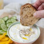 A bowl of whipped feta dip topped with olive oil and surrounded by colorful veggies. A piece of whole wheat sourdough bread is being dipped into the bowl of dip.