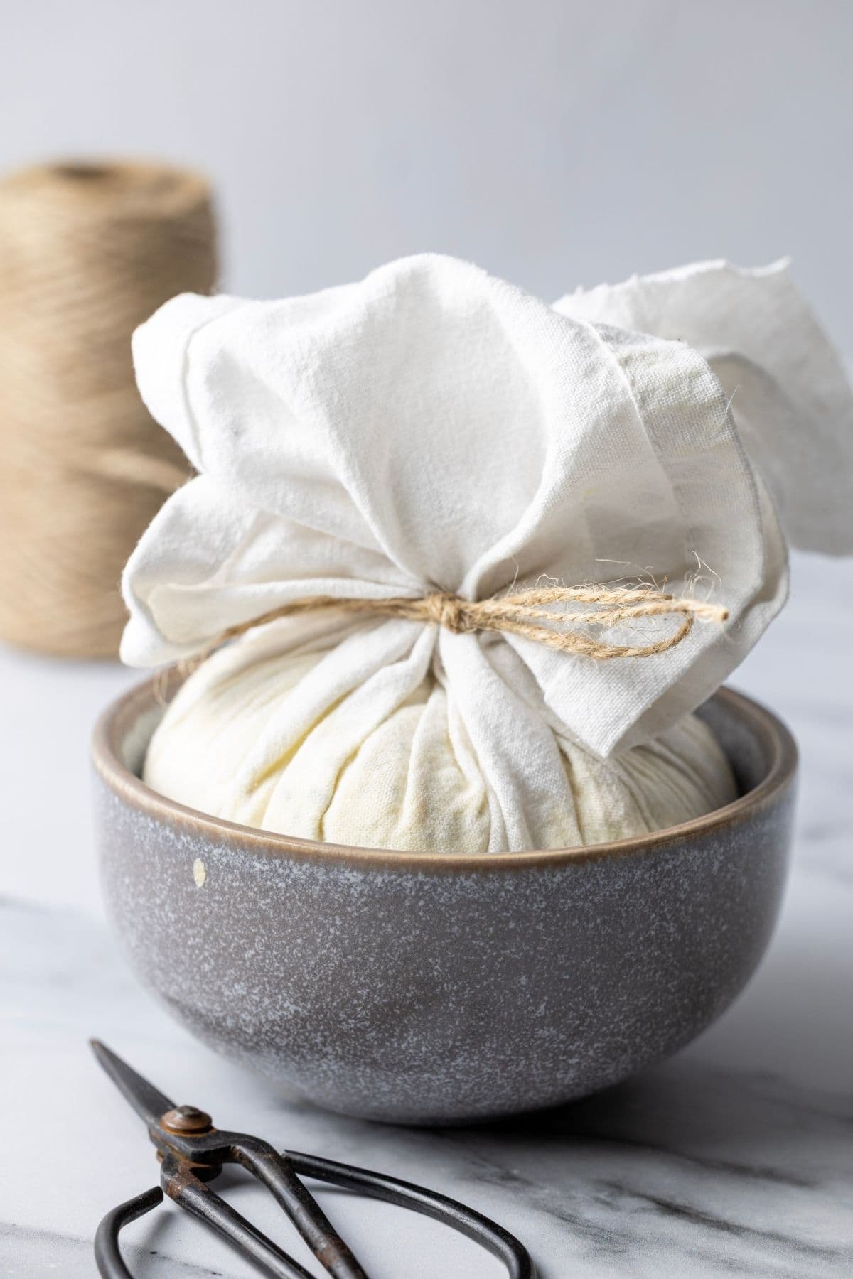 Homemade Boursin cheese wrapped in muslin and tied with twine, sitting in a grey stoneware bowl so that it molds to that shape. There are a small pair of antique scissors in the photo too.