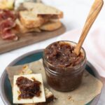 Jar of caramelized onion relish on a parchment paper lined plate next to a piece of sourdough bread topped with cheese and onion relish on top.