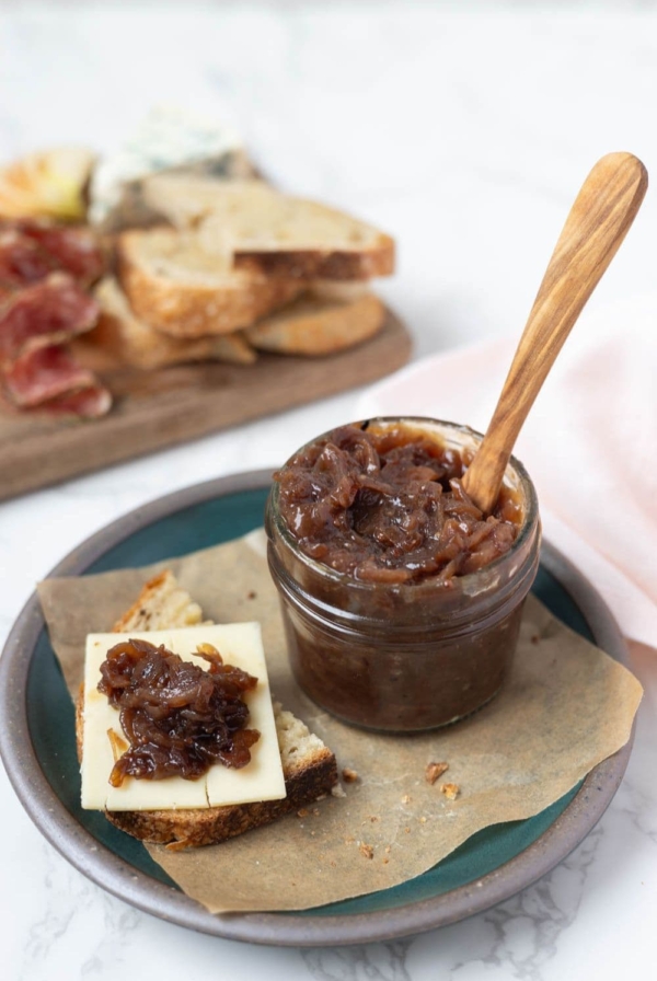 Jar of caramelized onion relish on a parchment paper lined plate next to a piece of sourdough bread topped with cheese and onion relish on top.