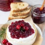 A wheel of Brie topped with vibrant cranberry vanilla jam. The Brie is decorated with fresh cranberries and rosemary and you can see the jar of jam and stack of sourdough bread slices in the background.