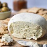 A block of homemade Boursin cheese that has been molded into a round shape and served with whole wheat sourdough crackers. The front of the cheese has been sliced open so you can see the texture inside. There is also a bowl of olives and jar of tomato relish in the background of the photo.