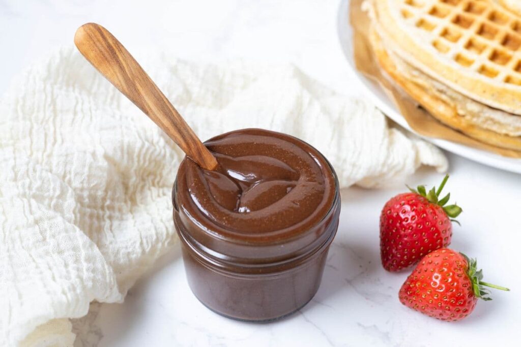 A jar of fresh made homemade Nutella with a wooden spoon inside, ready for adding to sourdough waffles.