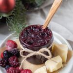 A small jar of mulled berry jam tied with a twine bow. The jar of jam is sitting on a plate alongside a selection of mixed berries and crumbly cheddar cheese. There is a Christmas wreath in the background of the jar.