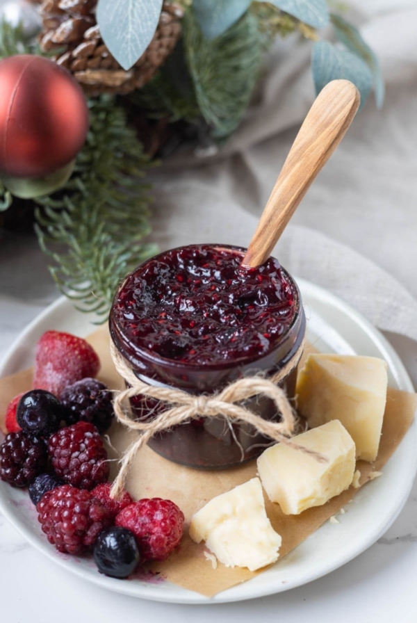 A small jar of mulled berry jam tied with a twine bow. The jar of jam is sitting on a plate alongside a selection of mixed berries and crumbly cheddar cheese. There is a Christmas wreath in the background of the jar.