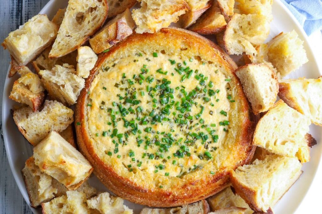 A sourdough cheese and garlic cobb loaf dip topped with fresh herbs and surrounded by toasted bread pieces for dipping.