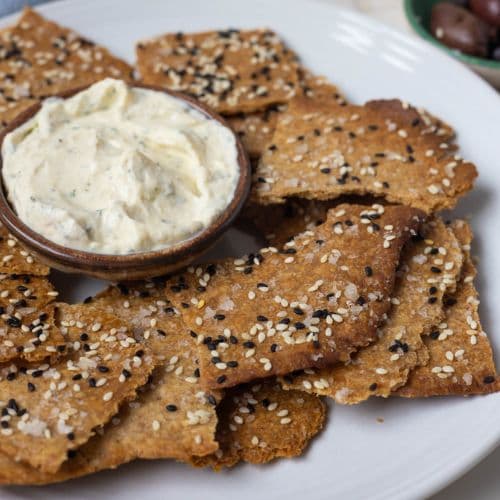 A selection of sourdough lavosh crackers topped with black and white sesame seeds surrounding a small bowl of dip.