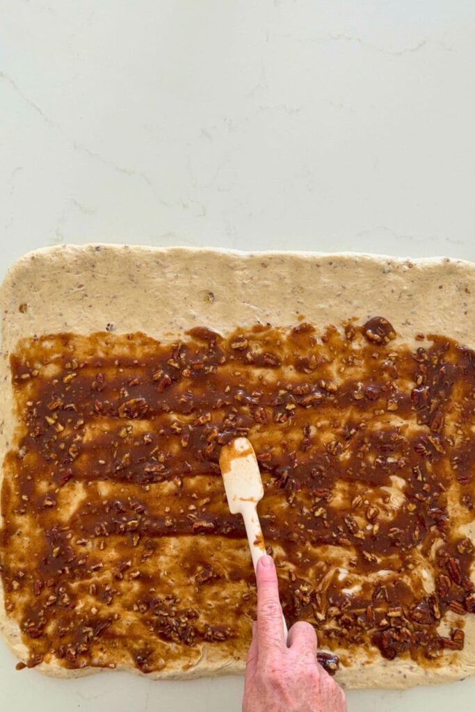 Dough for a sourdough maple pecan twist loaf stretched out on counter with a hand using a rubber spatula to spread filling over dough.