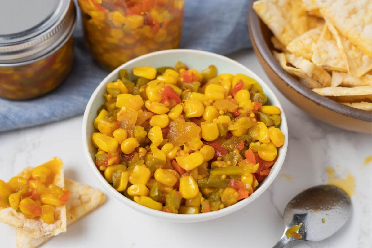 A bowl of corn relish on the counter with a bowl of chips and a spoon for serving.