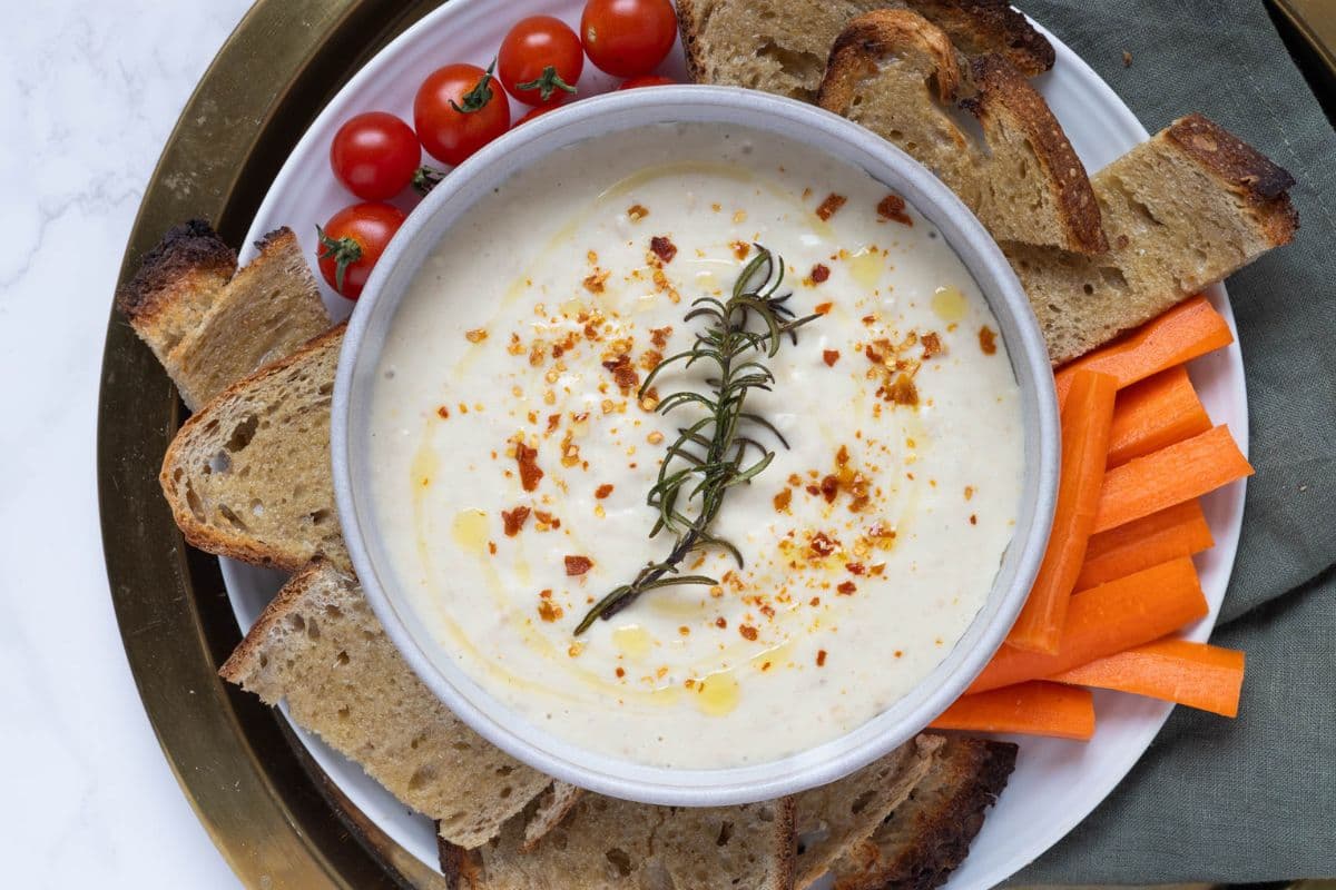 A serving plate featuring white bean dip topped with chili flakes and a toasted rosemary sprig on a platter with vegetables and sourdough toast pieces.