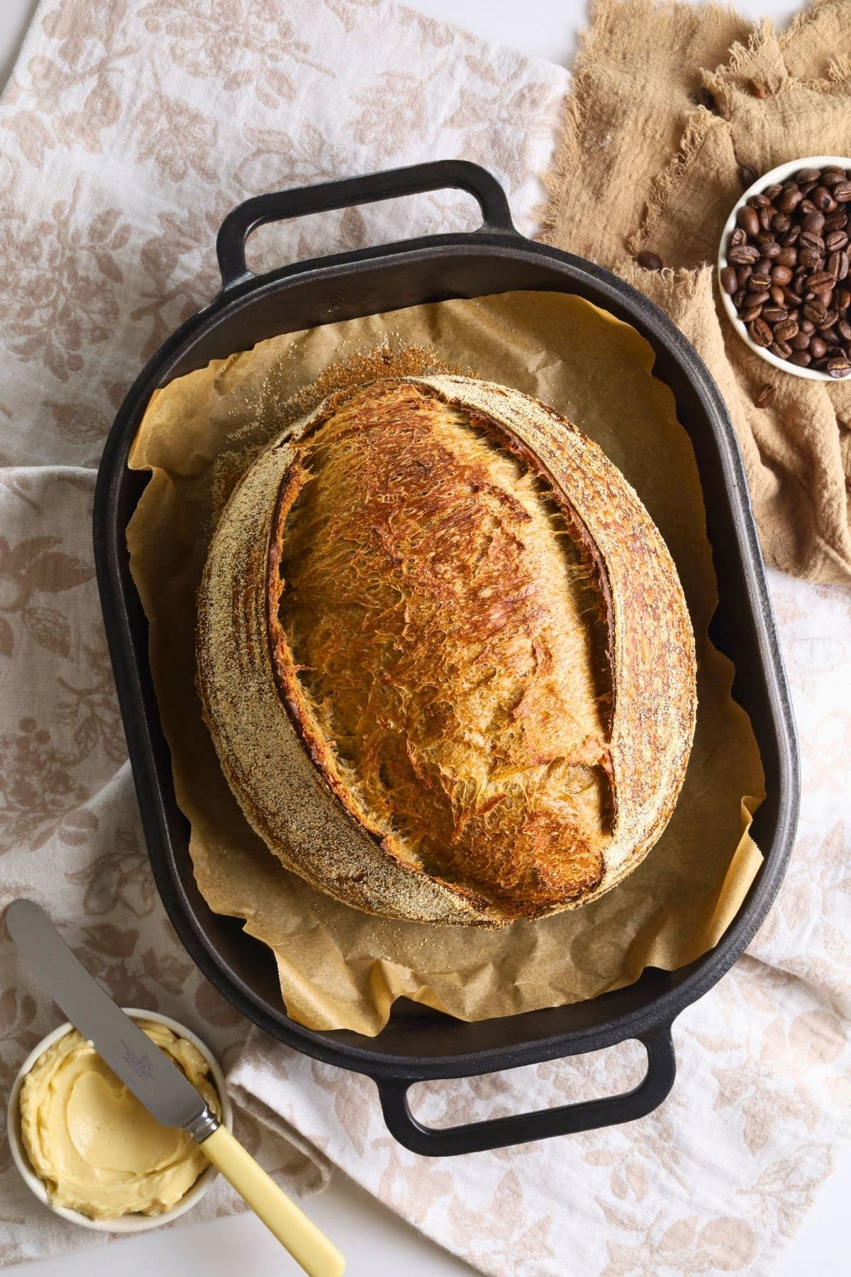 A loaf of sourdough bread made with cold brew coffee displayed in a Dutch Oven and photographed from above. There is a small dish of butter and vintage knife to the left of the image.