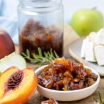 A small white dish filled with fresh peach chutney. The dish is flanked by a fresh peach half and the jar of peach chutney can be seen in the background. There is also a wheel of Brie and some rosemary sprigs, as well as a green apple in the very background.