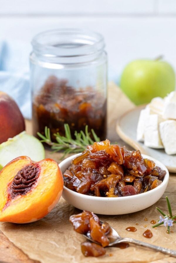A small white dish filled with fresh peach chutney. The dish is flanked by a fresh peach half and the jar of peach chutney can be seen in the background. There is also a wheel of Brie and some rosemary sprigs, as well as a green apple in the very background.