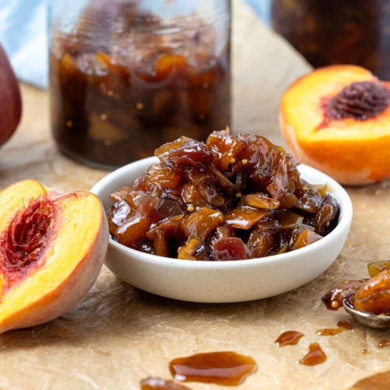 A small white dish filled with fresh peach chutney. The dish is flanked by 2 fresh peach halves and the jar of peach chutney can be seen in the background.
