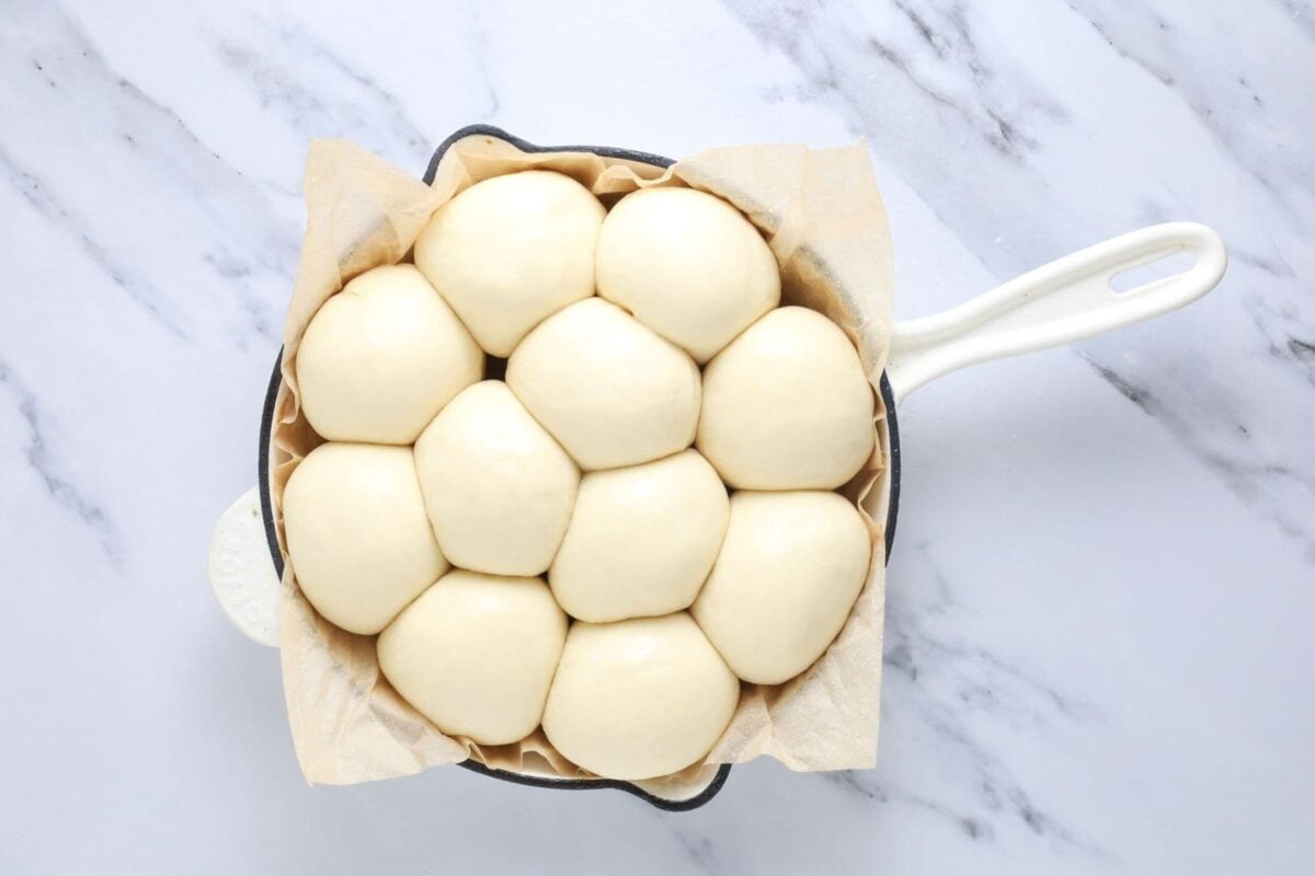 A photo showing sourdough discard rolls that have proofed inside a cream colored cast iron skillet. The rolls are soft and puffy and very squished together signalling that they are ready to be baked.