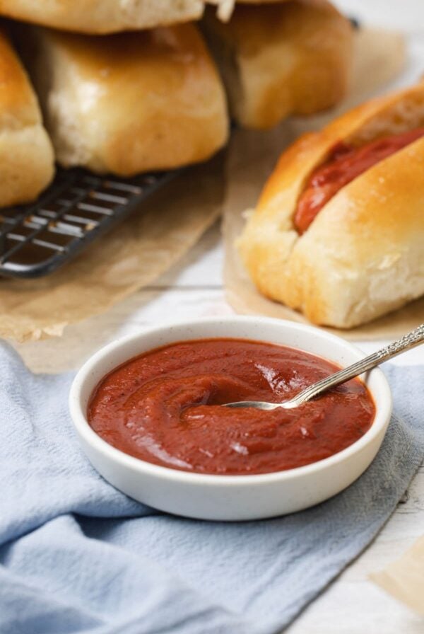 A bowl of homemade ketchup on the table with hot dogs for serving.