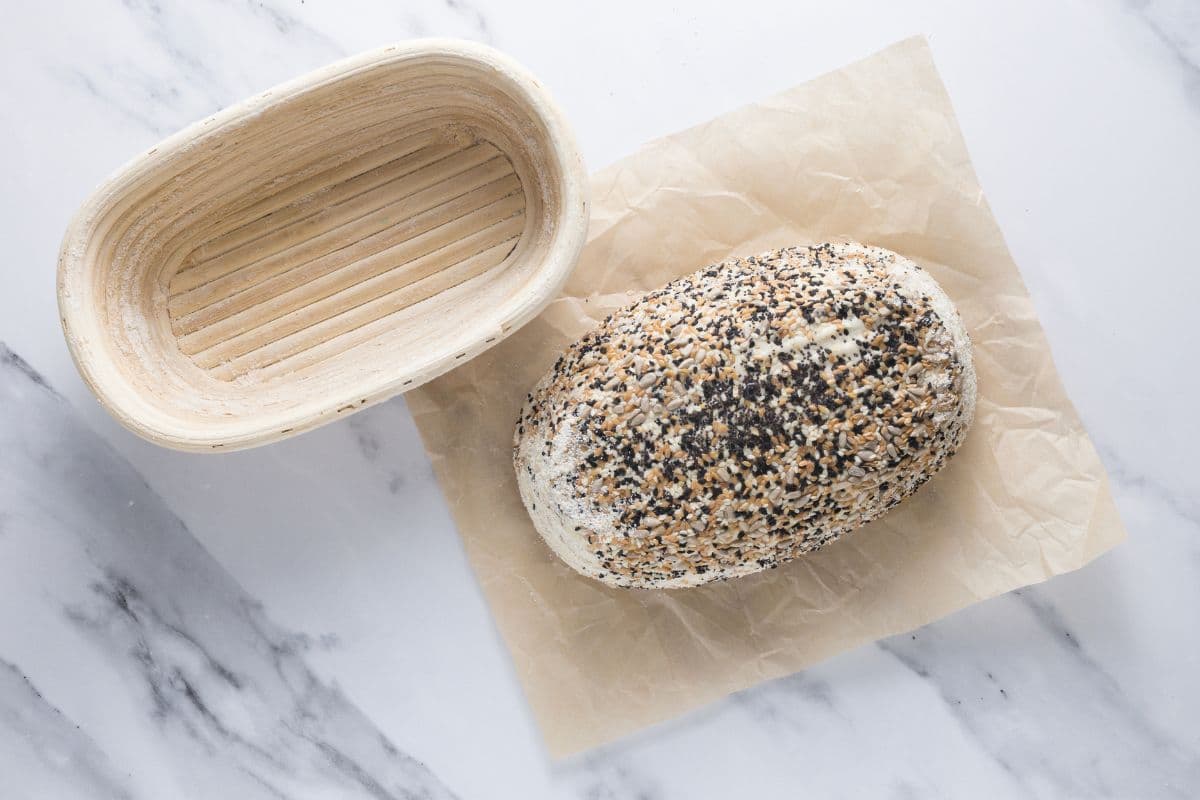 Shaped multigrain sourdough bread dough on a piece of parchment paper, next to a banneton after bulk ferment.