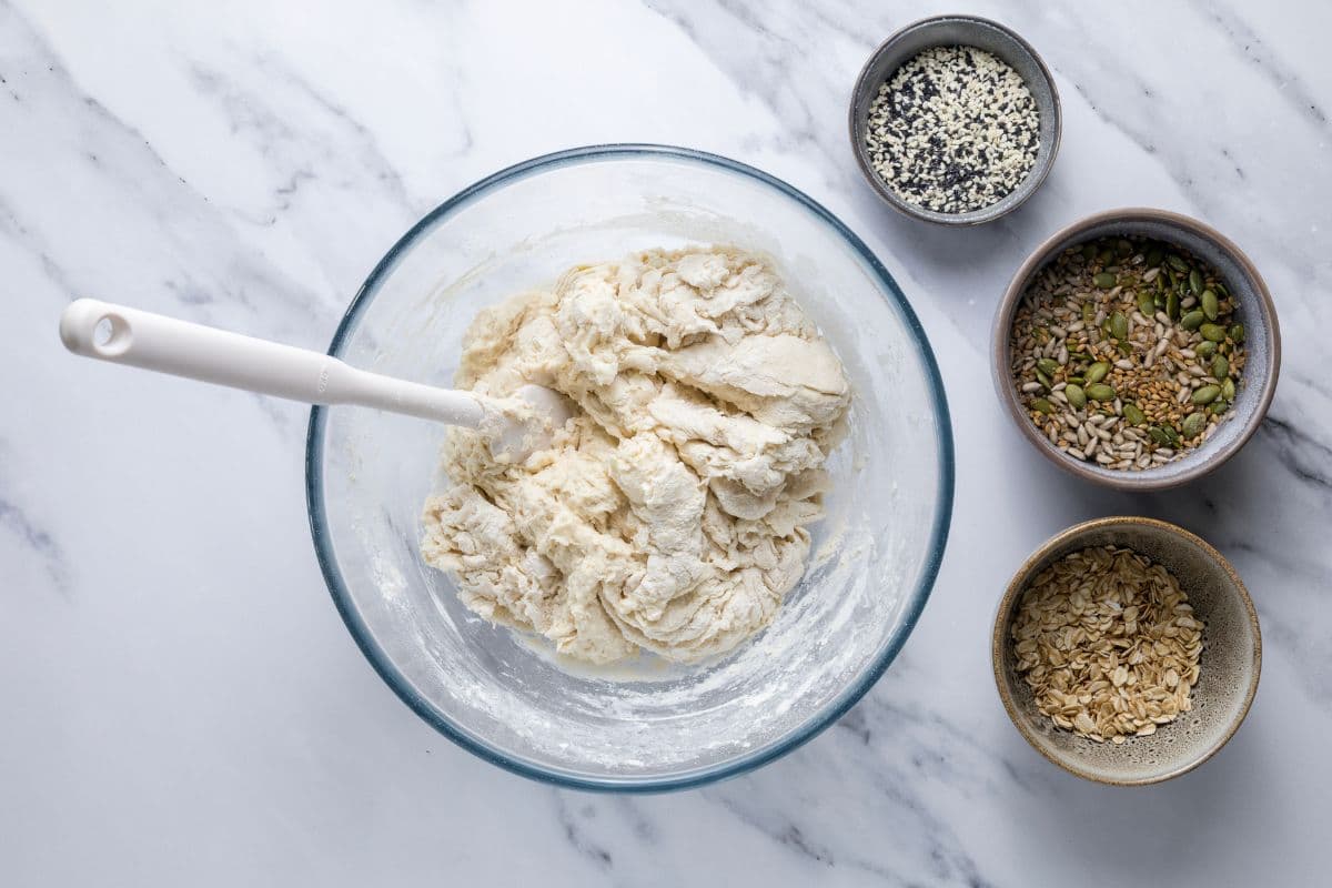 A bowl of rough mixed dough next to grains and seeds to make multigrain sourdough bread.