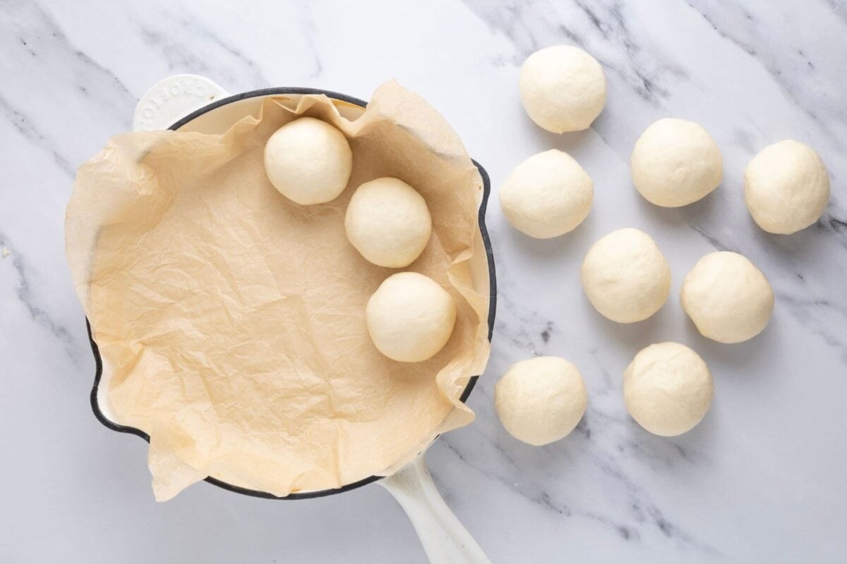 A photo of a parchment paper lined cast iron skillet. There are three dough balls inside the skillet and another 8 dough balls on the right hand side of the skillet.
