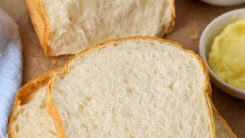 A loaf of sourdough discard sandwich bread that has been sliced up. Some of the slices are stacked on a wooden board with a small dish of butter and jam.