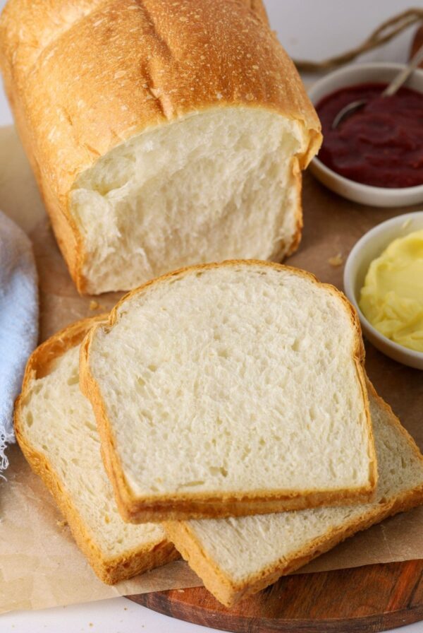 A loaf of sourdough discard sandwich bread that has been sliced up. Some of the slices are stacked on a wooden board with a small dish of butter and jam.