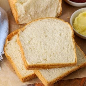 A loaf of sourdough discard sandwich bread that has been sliced up. Some of the slices are stacked on a wooden board with a small dish of butter and jam. You can also see the rest of the loaf in the background of the image.