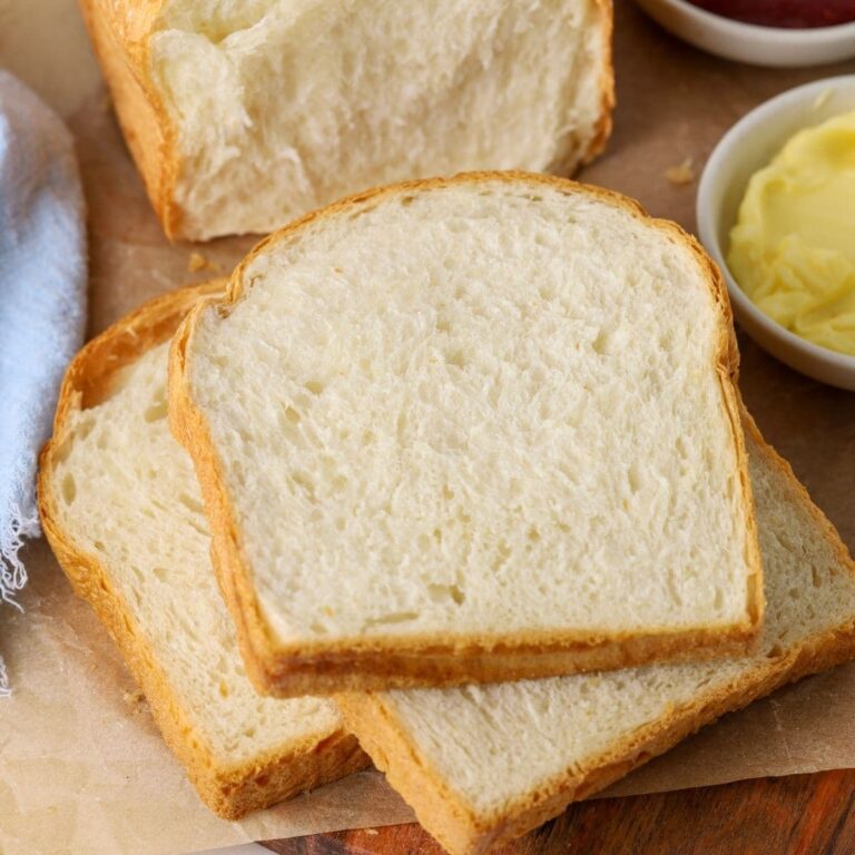 A loaf of sourdough discard sandwich bread that has been sliced up. Some of the slices are stacked on a wooden board with a small dish of butter and jam. You can also see the rest of the loaf in the background of the image.