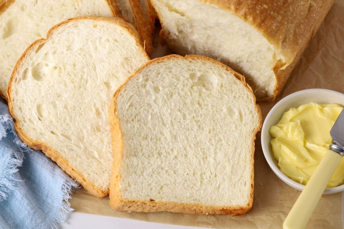 A loaf of sourdough discard sandwich bread that has been sliced up. Some of the slices are stacked on a wooden board with a small dish of butter and jam.