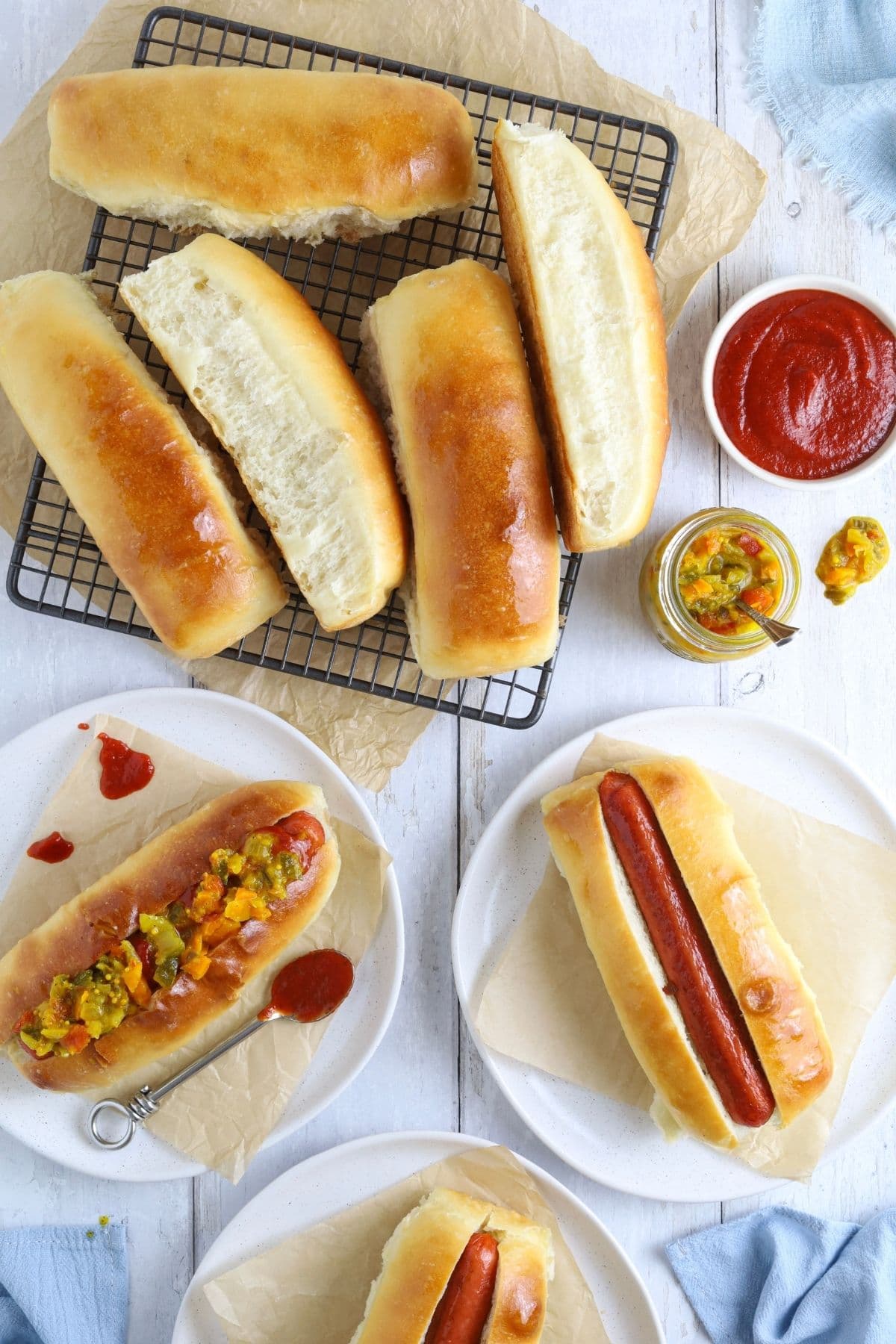 A flat lay image that has been taken from the top down. There are 6 freshly baked sourdough hot dog rolls in the top left corner, as well as 3 hot dog rolls filled with frankfurts and ketchup. There are two small bowls filled with ketchup and mustard relish in the picture too.