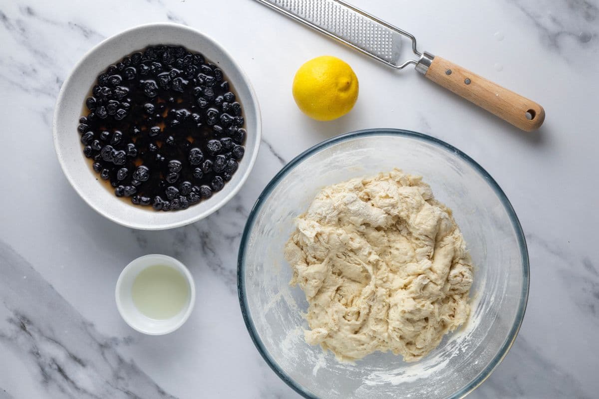 A bowl of blueberries soaking in maple syrup and a mix of shaggy dough to make sourdough lemon blueberry bread.