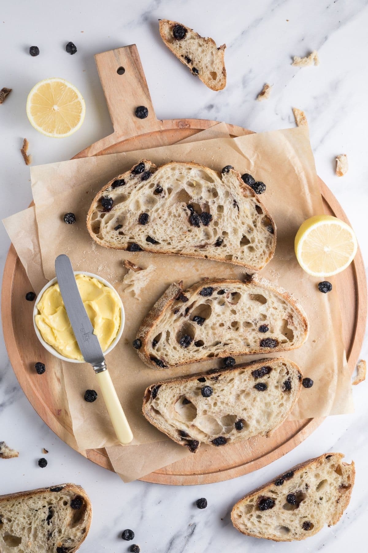 Slices of sourdough lemon blueberry bread on a parchment lined cutting board with a bowl of butter for serving.