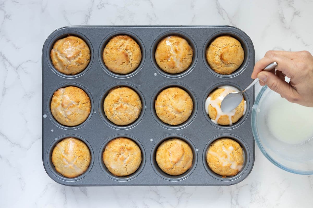 Lemon glaze being spooned over freshly baked sourdough lemon muffins in a muffin pan.