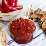 A close up of a jar of fresh chili jam being served with breads and a bowl of chili peppers.