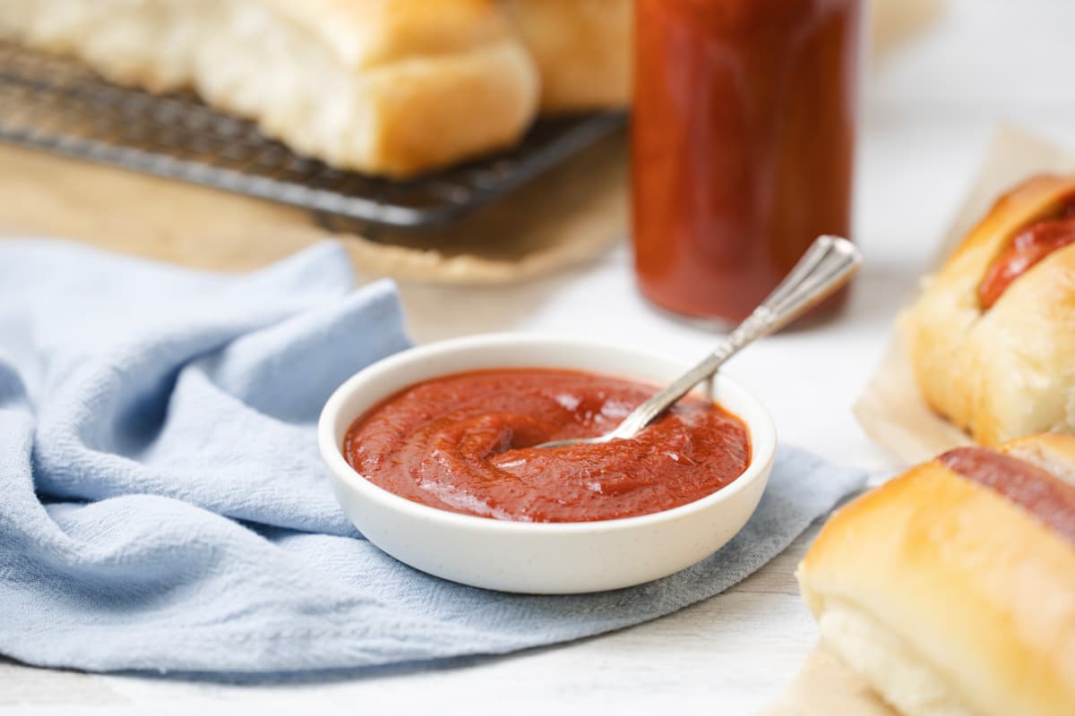 A bowl of homemade ketchup on a linen napkin being served with hotdogs.
