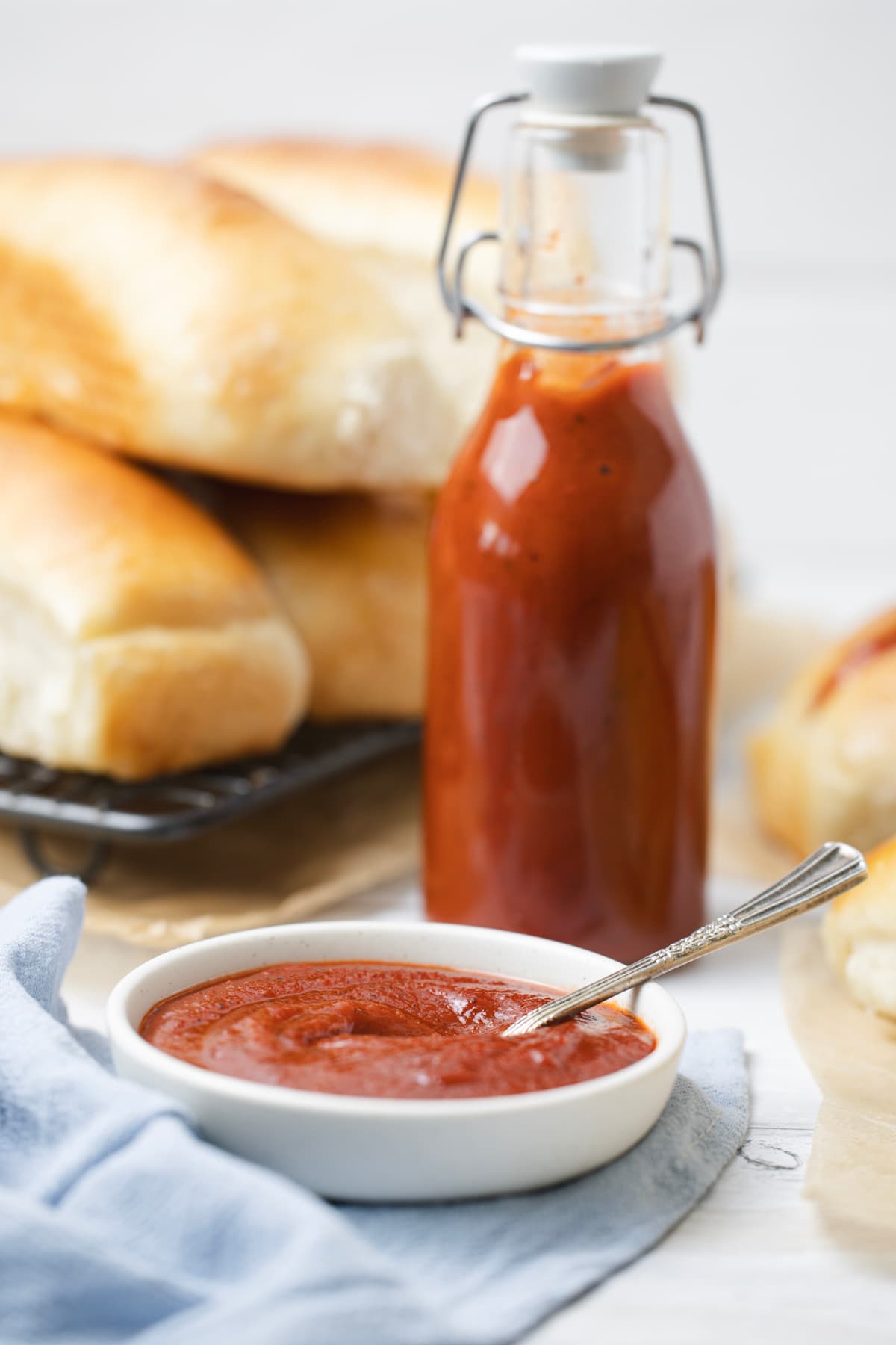 A bowl of homemade ketchup with a spoon in front of a bottle of homemade ketchup and a stack of hot dog buns.
