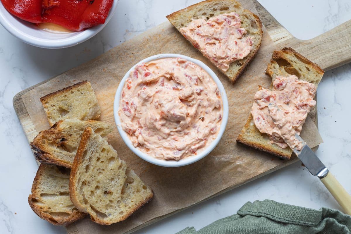 Roasted red pepper cream cheese dip served on a wooden board with sliced sourdough and roasted peppers.