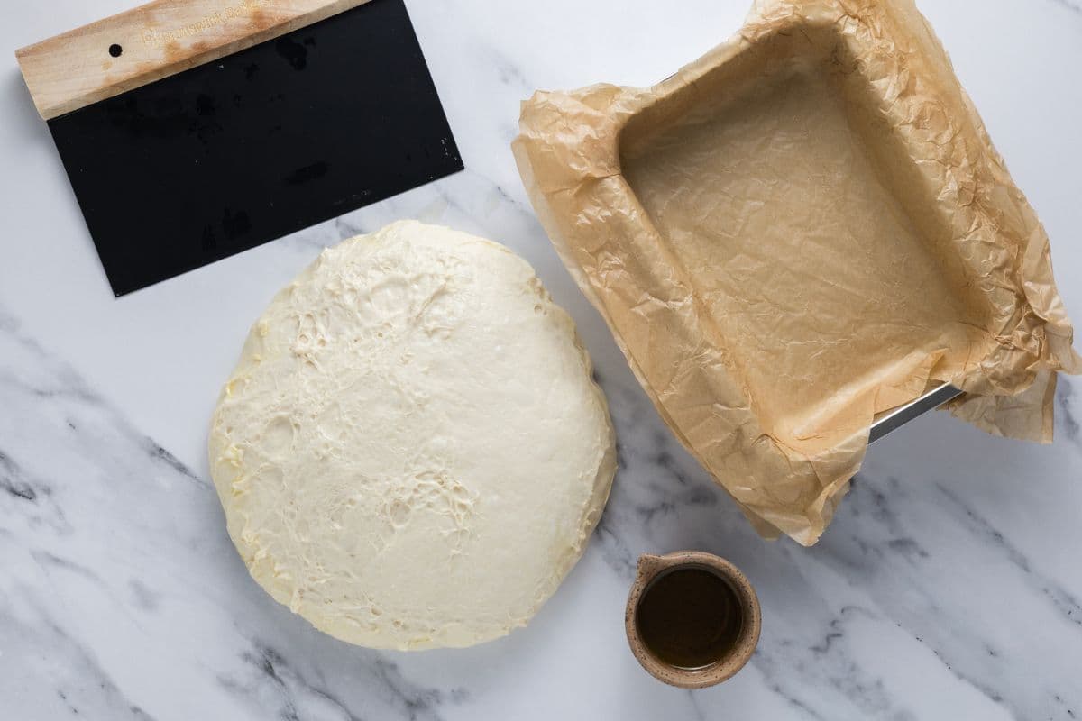 Adding simple sourdough focaccia bread dough into a parchment lined baking pan.