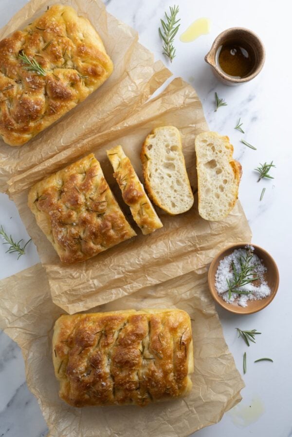 Three loaves of simple sourdough focaccia bread on parchment paper with one loaf sliced open to show inside texture.