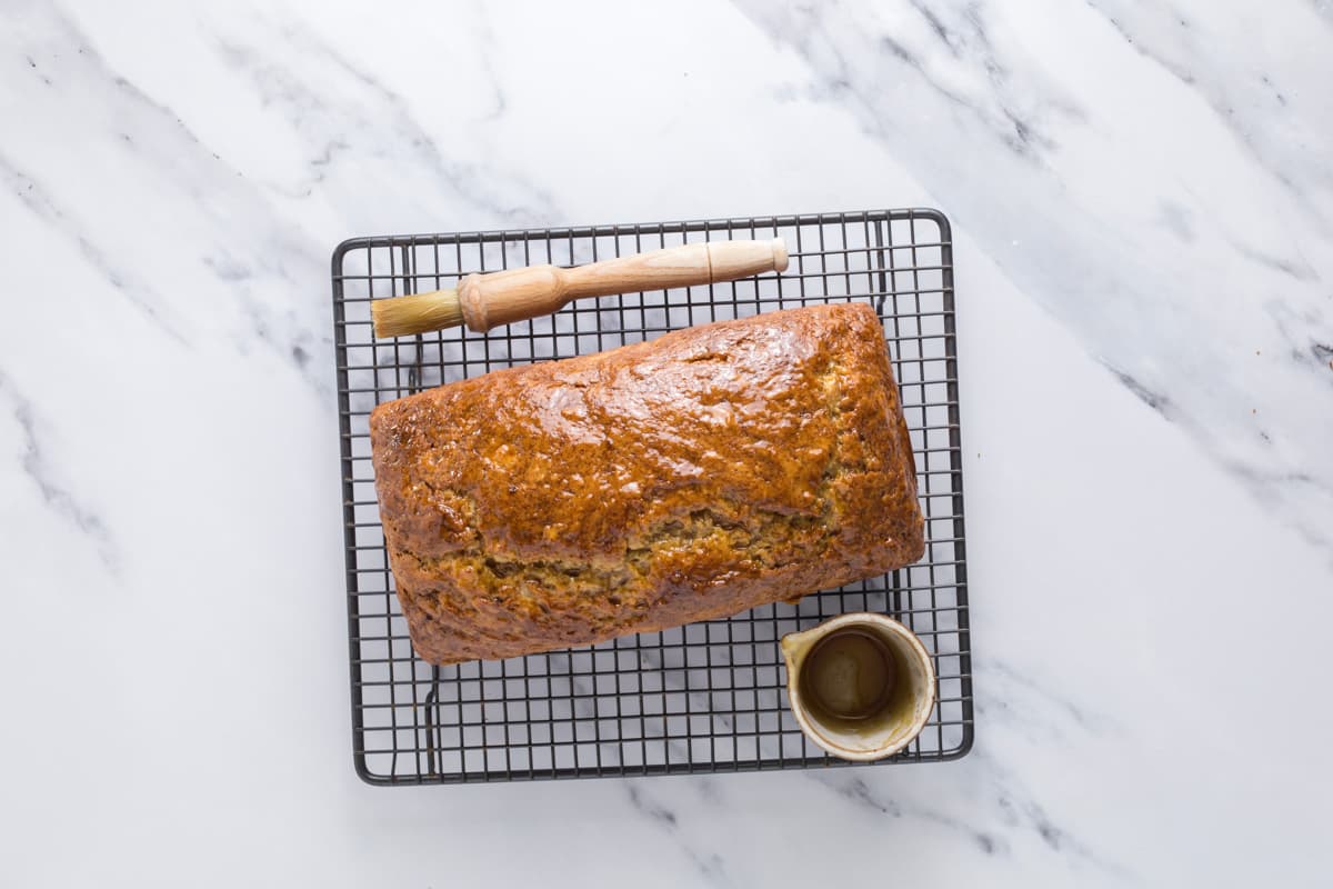 A fresh baked loaf of sourdough discard banana bread cooling on a wire rack just after being brushed with golden syrup.